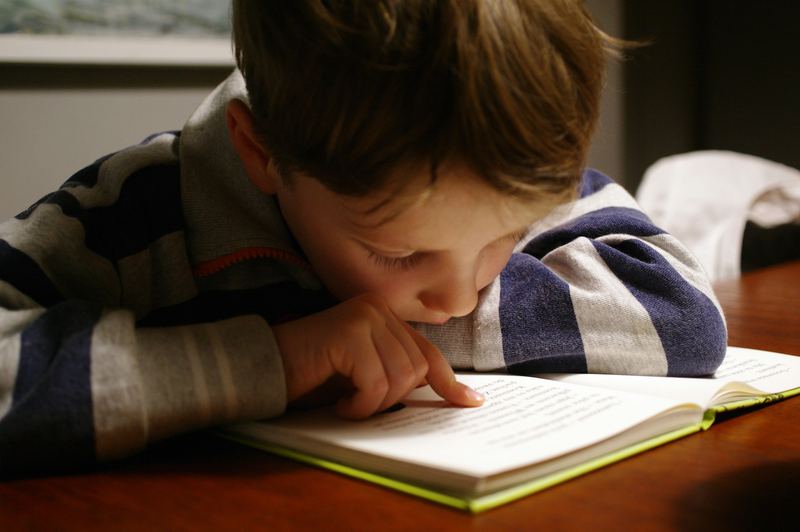 Menino lendo com o rosto bem perto do livro, acompanhando a linha com o dedo